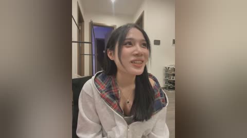 Media: Video of a smiling East Asian woman with long black hair, wearing a white jacket with a red plaid lining, standing in a narrow hallway with beige walls and a black couch visible in the background.