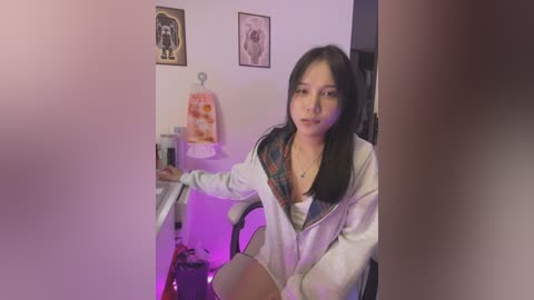 Media: Video of an Asian woman with long black hair, wearing a white jacket over a plaid shirt, sitting at a desk in a room with purple lighting, framed artwork on the wall, and a hanging towel.