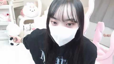 Media: A video of a young Asian woman with long black hair, wearing a white surgical mask, black hoodie, and pink gloves, in a brightly lit room filled with plush toys, including a white bunny.