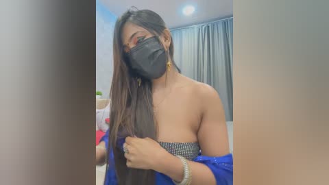 Media: A video of an Asian woman with long black hair, wearing a black face mask, a blue off-shoulder dress, and a beaded necklace, posing indoors with a blurred background.