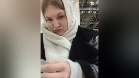 Media: Video of a young woman with light skin and light brown hair, wearing a white fluffy hooded robe, intently examining a piece of paper, set against a dimly lit, industrial background with visible beams and lights.