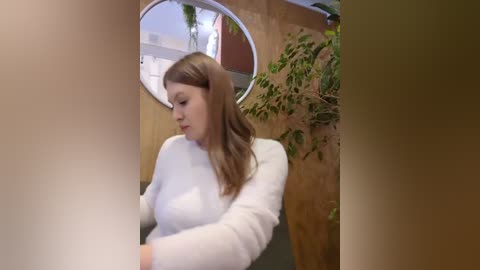 Media: A video of a young Caucasian woman with long, wavy brown hair, wearing a white sweater, standing in front of a round mirror and a green potted plant, in a modern, indoor setting.