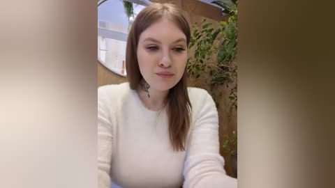 Media: Video of a young Caucasian woman with straight, shoulder-length brown hair, wearing a white long-sleeved shirt, standing outdoors against a wooden fence, looking contemplative.