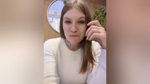 Media: Video of a young woman with fair skin, straight brown hair, and a white sweater, leaning on a wooden table with a circular mirror in the background.