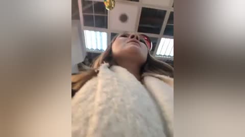 Media: Video of a young woman lying on her back in a room with fluorescent lights and white walls. Her face is tilted upwards, eyes closed, and she is wearing a white blanket.