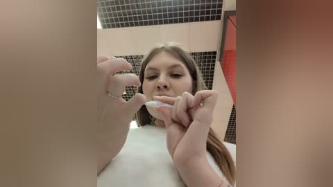 Media: Video of a young woman with light skin and long brown hair brushing her teeth in a bathroom with black and white tiled walls and a red towel.