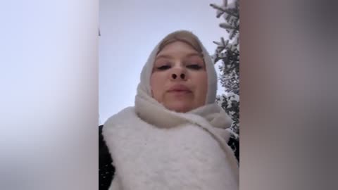 Media: Video of a blonde woman wearing a white woolen scarf, hood, and black coat, looking up with a neutral expression, surrounded by snow-covered trees in a winter landscape.