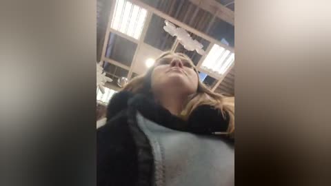 Media: A video of a woman with light brown hair, wearing a dark jacket and white shirt, taken from a low angle, inside a rustic, dimly lit room with wooden beams and fluorescent lighting.