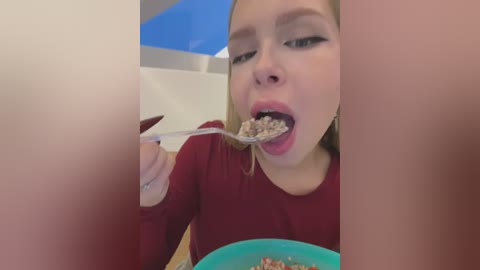 A video of a young woman with fair skin and blonde hair, wearing a red long-sleeve top, eating cereal from a blue bowl. The background shows a kitchen with white tiles and a blue wall.