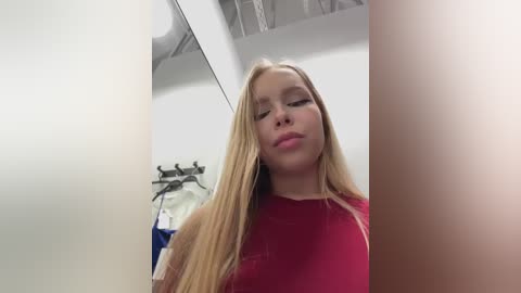 Video of a young blonde woman with fair skin, wearing a red shirt, standing in a brightly lit room with white walls, a black coat rack, and hanging clothes in the background.