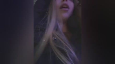 Media: Video of a woman with long, straight, blonde hair, wearing a black leather jacket, standing in a dimly lit room with a dark background, slightly blurred, focusing on her face and shoulders.