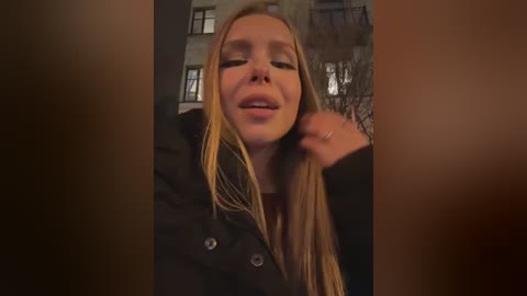 Video of a young Caucasian woman with long blonde hair, wearing a black coat, standing in a dimly lit urban setting with blurry apartment buildings and a metal railing in the background.