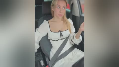 Media: Video of a blonde woman with fair skin, wearing a white dress with a black ribbon, sitting in a car, looking pensive.