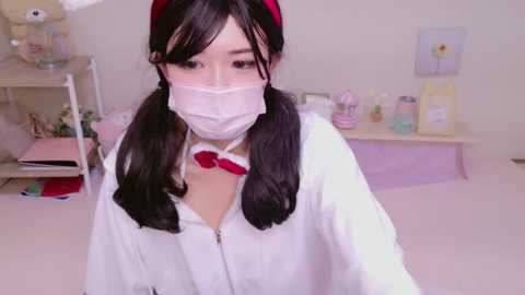 Media: A young Asian woman with long black hair in pigtails, wearing a white medical mask, red headband, and white lab coat, takes a selfie in a pink-tiled room with medical supplies and a shelf.