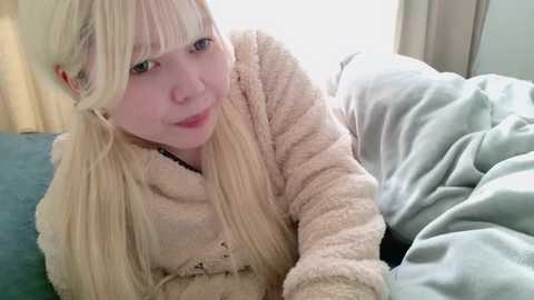 Media: Video of a fair-skinned woman with long, straight blonde hair, wearing a beige fleece robe, lying on a bed with white sheets and a teal blanket. The background includes a white curtain and a soft, beige carpet.