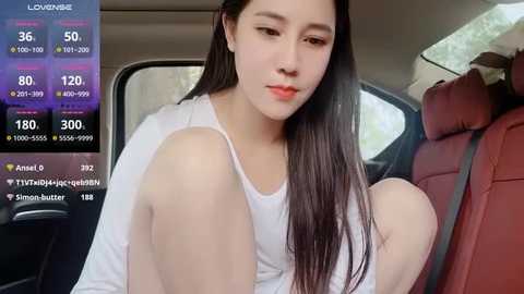 Media: A video of a young East Asian woman with long black hair, fair skin, and red lipstick, wearing a white tank top, sitting in a red car seat.