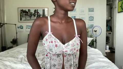 Media: Video of a dark-skinned woman with short hair, wearing a floral lingerie set, standing in a modern, minimalist bedroom with white walls, a bed, and various framed art.