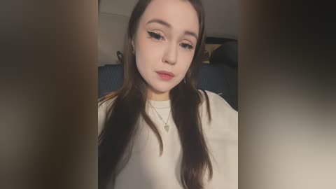 Video of a young woman with fair skin, long dark hair, and full makeup, wearing a white sweater, taken indoors with blurry background.