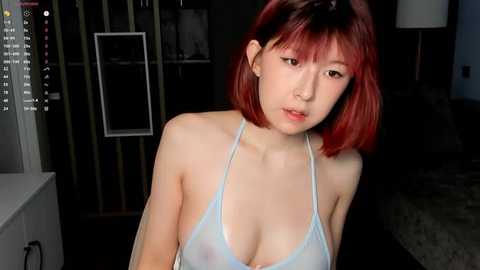 Media: Video of a young East Asian woman with short, red hair and pale skin, wearing a light blue, see-through spaghetti strap top, revealing her breasts. She gazes downwards in a dimly lit, modern room.