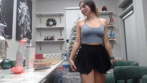 Media: Video of a young, fit woman with light skin and brown hair in a gray sports bra and high-waisted black shorts, standing in a cluttered, modern kitchen with white cabinets, green chairs, and a Christmas tree.