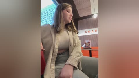 Media: A video of a young, fair-skinned woman with shoulder-length blonde hair, wearing a beige cardigan and a light-colored top, sitting in a modern office with wooden accents and a \"NIDA\" sign.