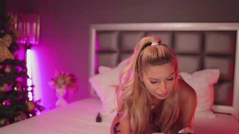 Media: Video of a blonde woman with long hair in a ponytail, wearing a black top, leaning over a bed in a dimly lit room with purple and pink lighting.