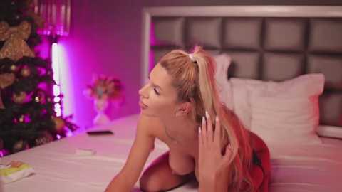 Media: Video of a blonde woman with a high ponytail, topless, leaning over a bed in a dimly lit bedroom with a Christmas tree and purple lighting.