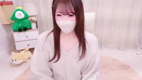 Media: Video of a young Asian woman with long brown hair, wearing a white face mask and a white hoodie, sitting on a beige rug in a minimalist room with white walls and a green plush toy.