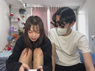 Media: Video of two young Asian women in casual attire, one with long brown hair, the other with short black hair, wearing masks, sitting on a bed in a cluttered room with shelves, toys, and pink curtains.