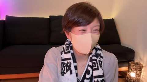 Media: Video of an Asian man with short, dark hair, wearing a beige face mask, a black and white patterned scarf, and a light-colored jacket, seated on a black couch in a dimly lit room with a small wicker lamp on the right.