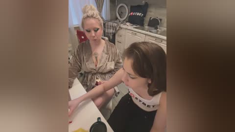 Media: A candid video of two women in a kitchen. The blonde, wearing a satin robe, holds a knife, while the brunette with a ponytail, in a black apron, watches intently.