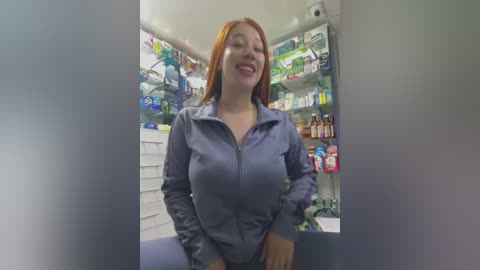 Video of a smiling Asian woman with shoulder-length red hair, wearing a blue zip-up jacket, standing in a well-lit pharmacy with shelves full of medicines.