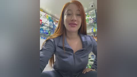 Video of a smiling, fair-skinned woman with long red hair, wearing a blue zip-up hoodie, seated in a brightly lit, well-stocked pharmacy aisle.