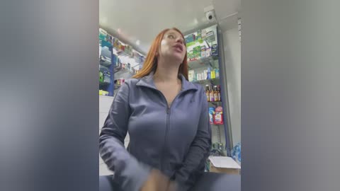 Video of a red-haired, fair-skinned woman in a blue jumpsuit, reaching into a brightly lit convenience store cooler filled with beverages.