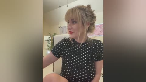 Media: Video of a blonde woman with a high ponytail, wearing a black polka-dotted dress, sitting in a modern office with beige walls, potted plants, and white chairs.