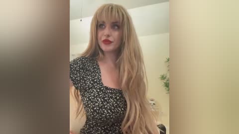 Media: Video of a fair-skinned, blonde woman with long, wavy hair wearing a black and white patterned dress. She has bold red lipstick and is standing in a beige room with a green plant in the background.