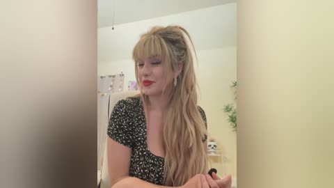 Media: Video of a blonde woman with long hair in a ponytail, wearing a black floral dress, smiling, standing in a cream-colored room with a medical cabinet and plants in the background.