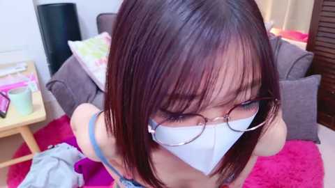 Video of an Asian woman with shoulder-length dark hair, wearing glasses and a white face mask, sitting on a pink rug in a cozy living room with a gray couch and wooden furniture.