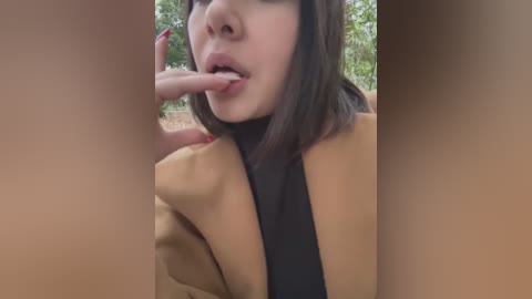 Media: Video of a woman with pale skin and straight brown hair, wearing a brown coat, licking a finger. Background shows blurred greenery.