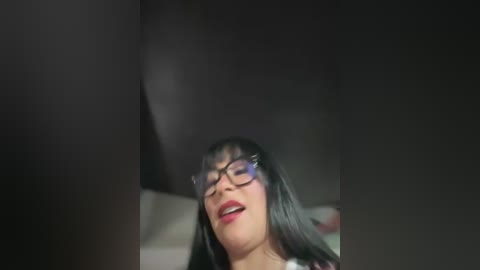 A video of a woman with long black hair, wearing glasses and red lipstick, looking up with a sensual expression. The background is dark and out of focus.