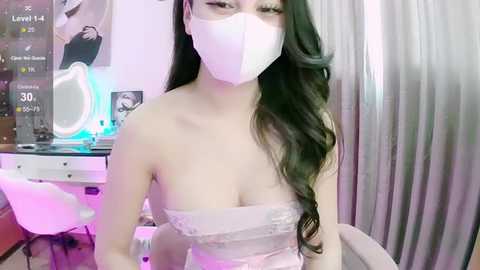 Media: A video of an Asian woman with long, wavy black hair, wearing a white face mask, a strapless white lace top, and sitting in a dimly lit room with a laptop, a makeup mirror, and a pink chair.