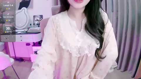 Media: Video of a young Asian woman with long black hair, wearing a frilly cream blouse, indoors. Background includes a vanity table with makeup, a white chair, and a grey curtain.