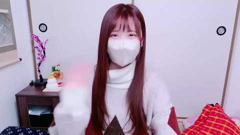 Media: Video of an East Asian woman with long, straight brown hair, wearing a white surgical mask and a red and white patterned sweater. She sits in a bedroom with white walls, a wooden nightstand, and a red plaid blanket.