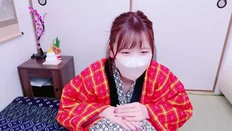 Media: Video of a young woman with a white face mask, wearing a red plaid sweater over a black-and-white patterned shirt, seated on a tatami mat in a traditional Japanese room with a wooden nightstand and floral decorations.