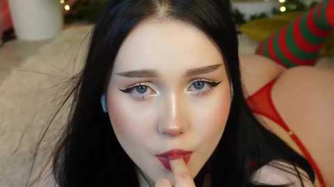 Media: Video of a pale-skinned woman with long black hair and bold black eyeliner, seductively licking her finger in red lingerie, with festive, blurred background.