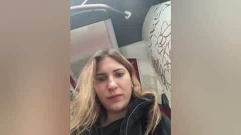 Media: Video of a young Caucasian woman with long, blonde hair, wearing a black jacket, seated in a train compartment. The background features a white wall with floral patterns and a black handle.