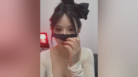 Media: Video of an East Asian woman with fair skin, wearing a black lace mask, white cardigan, and black hair in a bun, sitting indoors with a red lamp and white walls in the background.