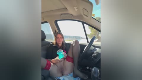 Media: A video of a woman with long hair, wearing a black top, red thigh-high stockings, and no pants, sitting in a car with her legs spread, holding a blue drink, in a sunny, scenic landscape.