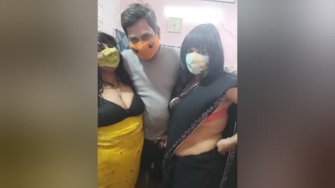 Media: Video of a man in a grey shirt, wearing a face mask, posing with two women in traditional Indian attire; one in a yellow blouse, the other in a black sari with a red blouse.