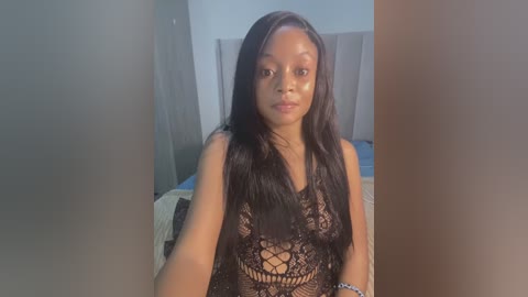 Media: Video of a young Black woman with long, straight black hair, wearing a black lace crop top, standing in a dimly lit bedroom with a white headboard and gray walls.